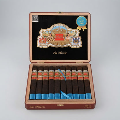 An open wooden cigar box on a light gray studio background, containing ten cigars. The interior lid is black with a large decorative La Historica crest and a blue cigar award seal.