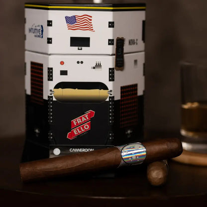 A cigar displayed in front of a branded storage box shaped like a spacecraft, with two cigars on a dark wooden surface and a glass of amber liquid in the background.