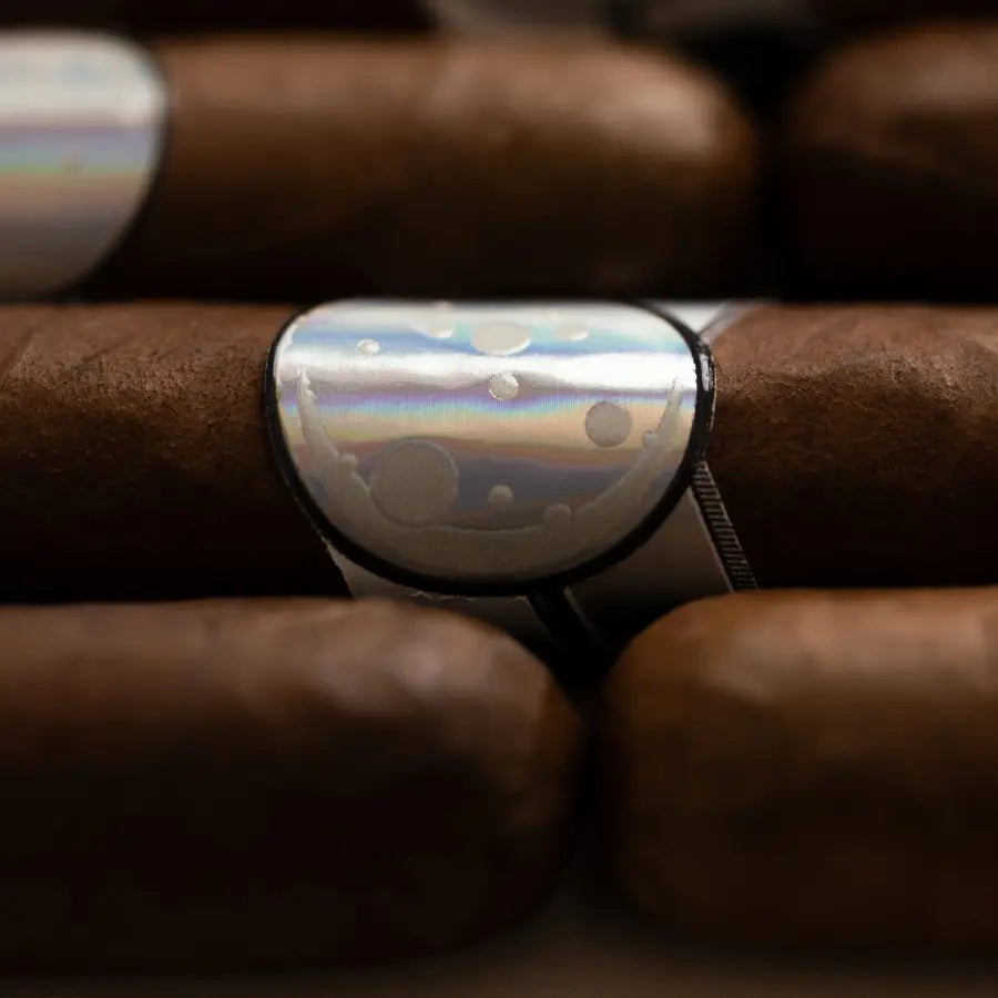 A close-up of a silver-toned ring with an iridescent, pearlescent face featuring white and gray cloud-like patterns, shown resting on a brown cigar.
