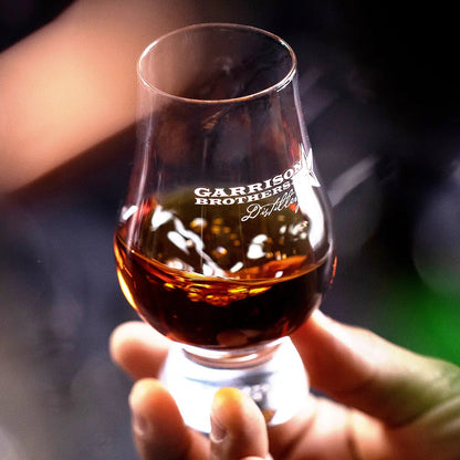 Hand holding a glass of whiskey with 'Garrison Brothers' branding against a blurred background
