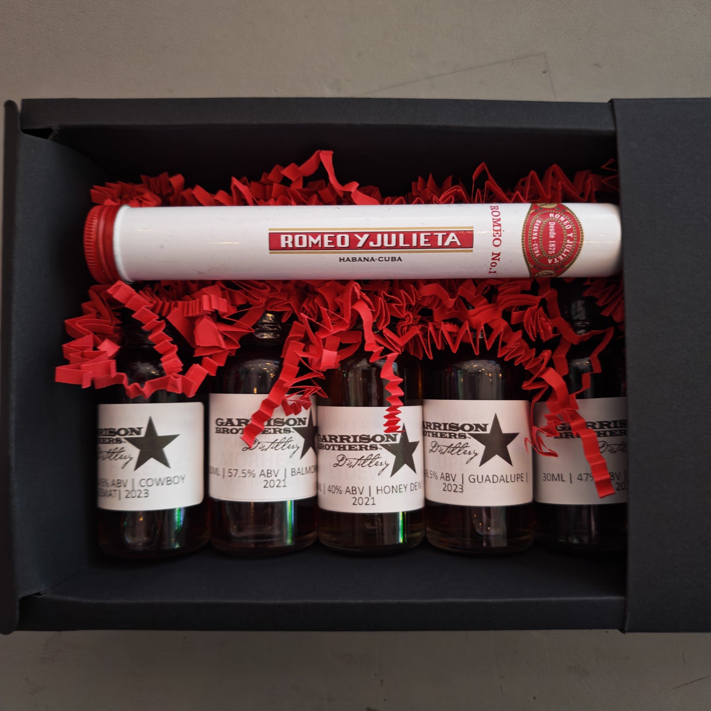 Set of miniature bottles of Garrison Brothers whiskey with labels and a tube labeled 'Romeo y Julieta' in a black box.