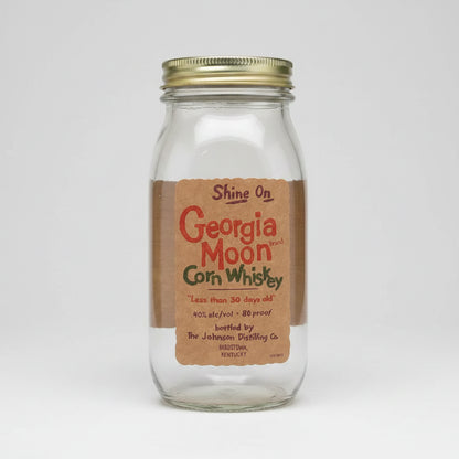 A clear glass jar with a metal screw-top lid containing a brown powder, featuring a paper label that reads 'Shine On Georgia Moon Brand Corn Whiskey' on a white background.