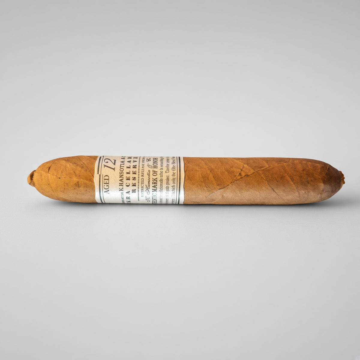 A single cigar shown horizontally against a light gray studio background. The cigar is medium brown with a white paper band around the middle that reads 'Aged 12' and includes the brand name 'Pabellon' and 'Cigarro de Pabellon'.