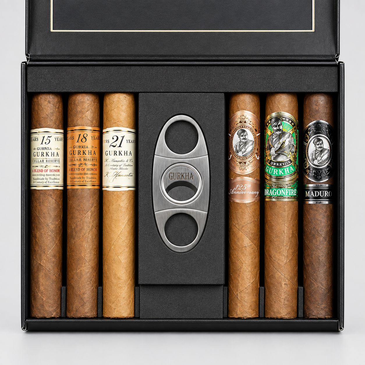 A premium cigar gift set in an open black presentation box, featuring eight cigars from the brand Gurkha in various shades of brown, a silver metal cigar cutter, and a black foam insert.