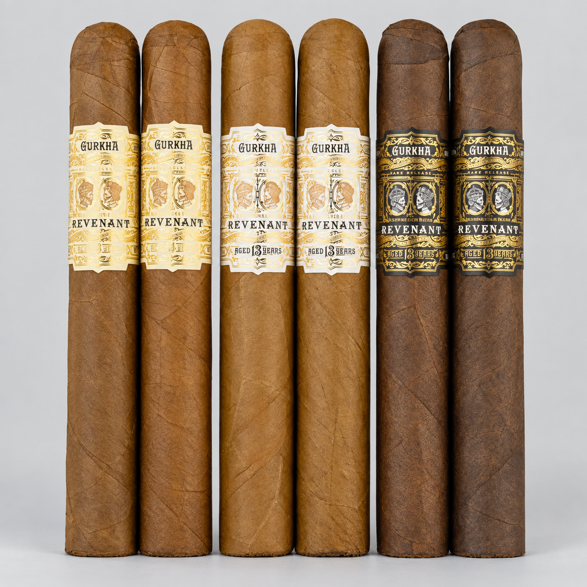 Six Gukha Revenant cigars arranged upright in two columns of three against a light gray studio background. The cigars show natural brown to dark brown leaf tones, with three cream-and-gold bands and three black-and-gold bands.