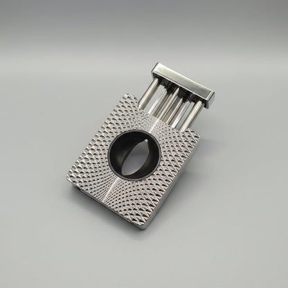 A silver metal cigar cutter with a textured diamond-patterned body and a black circular cutting ring, shown open on a brown wooden surface.