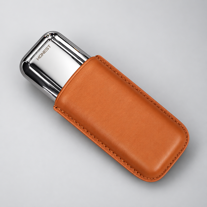 A silver metal personal care device, branded HONEST, nestled inside a tan leather protective sleeve with visible stitching, set against a clean light gray background.