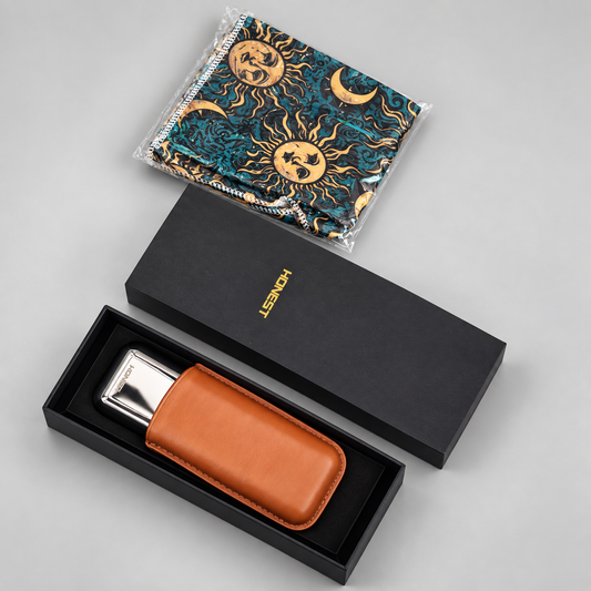 A silver rectangular metal accessory with a brown leather sleeve presented in an open black box, alongside a black branded box lid and a folded patterned cloth in plastic packaging on a light gray background.