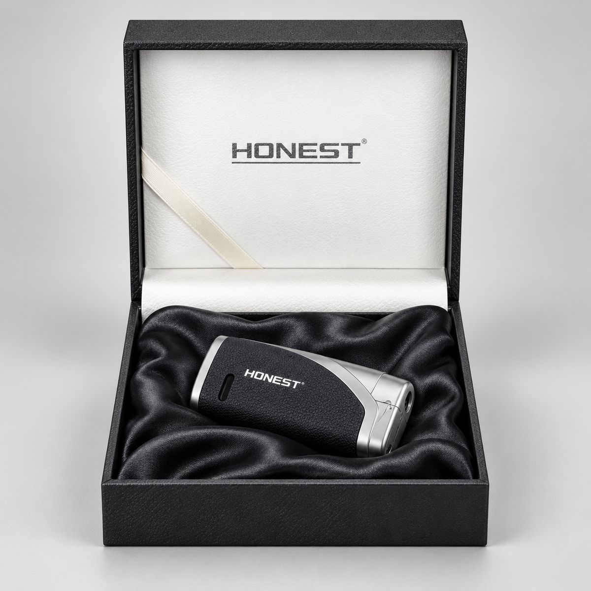 A black and silver Honest lighter resting on a black satin cushion inside an open black presentation box with a white interior and the Honest logo printed on the lid.