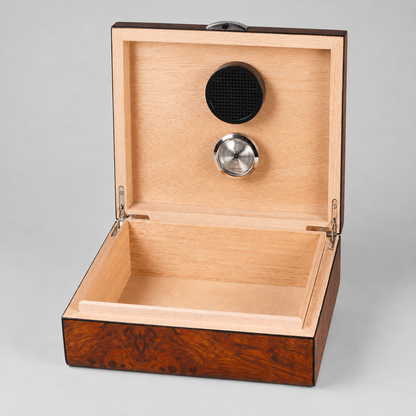 An open wooden presentation box with a light natural wood interior and a darker brown burl wood exterior. The inside lid features a black circular speaker and a silver metal button, set against a neutral gray background.