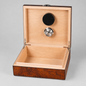 An open wooden presentation box with a light natural wood interior and a darker brown burl wood exterior. The inside lid features a black circular speaker and a silver metal button, set against a neutral gray background.