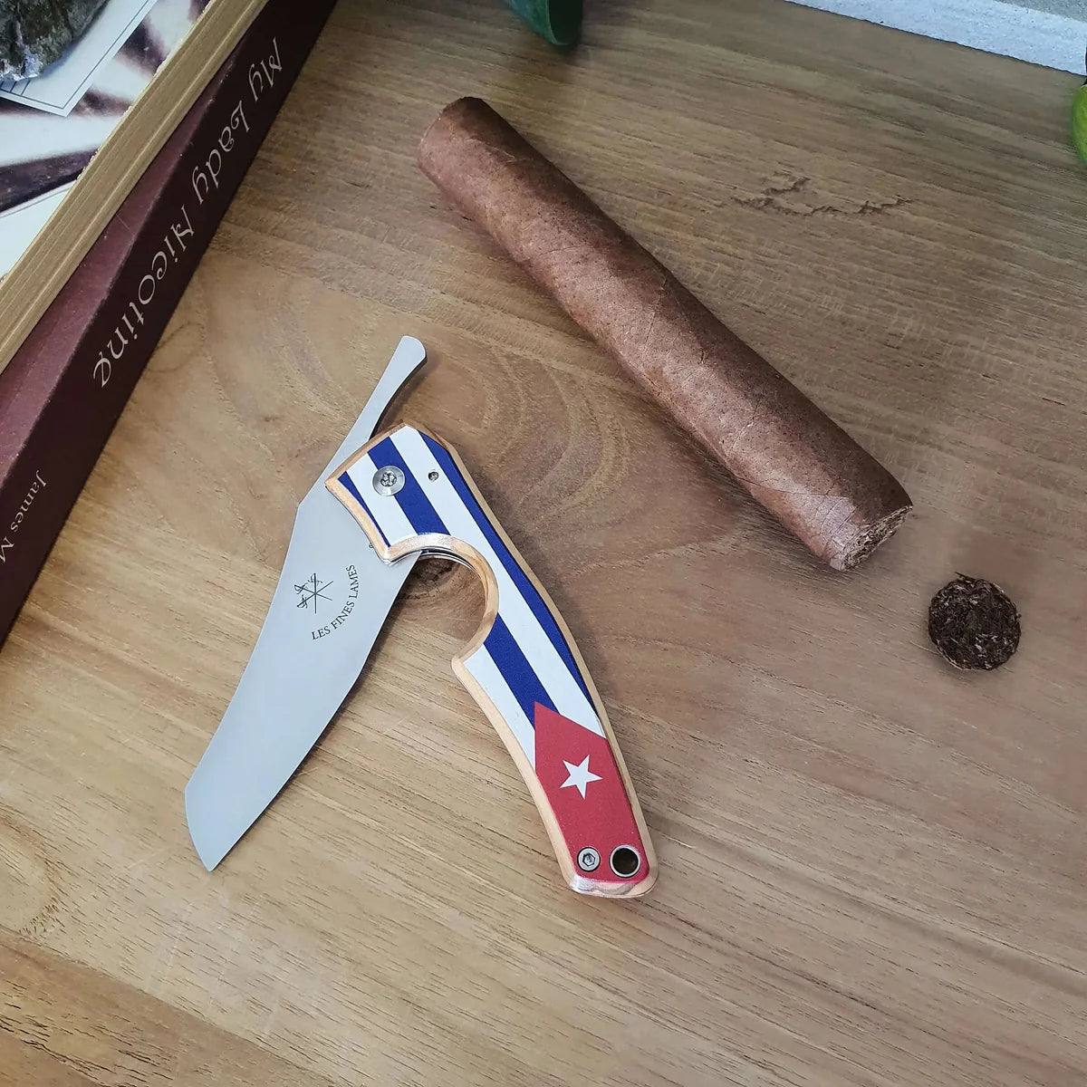 A folding pocket knife with a Cuban flag pattern handle lies on a wooden tabletop next to a cigar and a small cigar butt. The knife blade is open, showing the stainless steel blade and the handle design with red, white, and blue stripes and a white star.