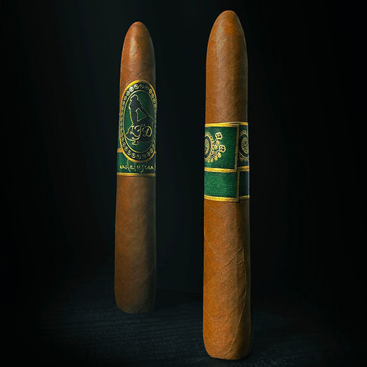 Two brown cigars standing upright against a dark black background, each with a green and gold band. The left band features a circular crest with 'Aguinaldo' and '1864' text, while the right band has a decorative green and gold pattern.