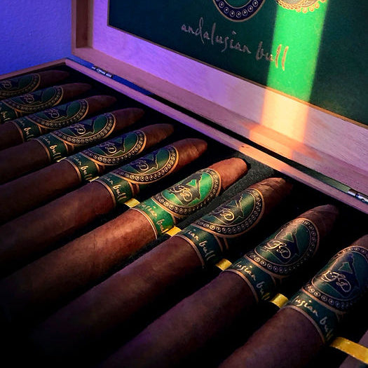 An open wooden cigar box containing several dark brown cigars with green and gold bands. The inside lid features a green panel with gold decorative graphics and the text 'andalusian bull'.