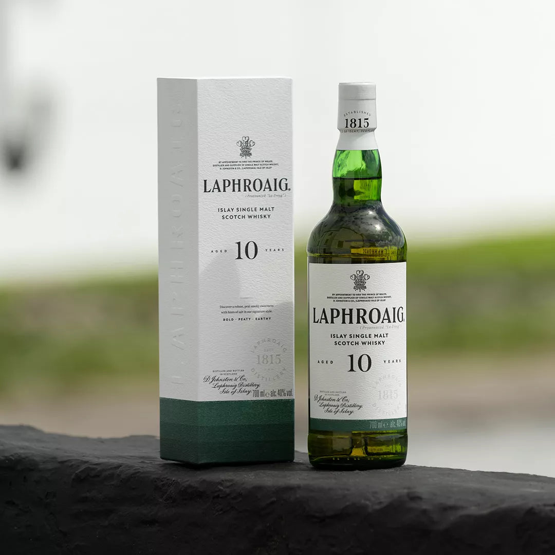A bottle of Laphroaig 10 Year Old single malt Scotch whisky stands next to its white and dark green packaging box on a dark stone surface, with a softly blurred outdoor background.