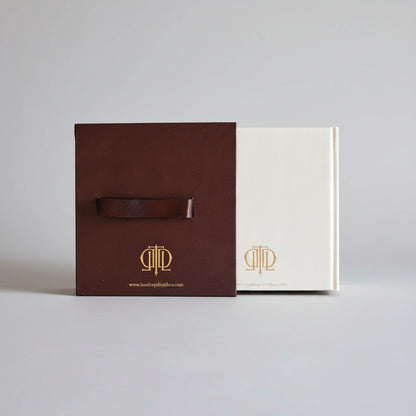 A brown leather strap with a gold logo attached to a dark brown rectangular box, placed on top of a cream-colored booklet or card, all on a textured blue background.