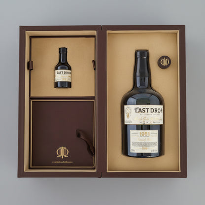 A whisky gift set from The Last Drop, shown in an open dark brown presentation box on a blue background. The set includes a small bottle, a larger bottle with a 1961 label, a round cap, and a dark brown box with a gold monogram logo.