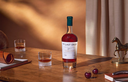Bottle of 'The Last Drop' whiskey on a wooden table with glasses and cherries.