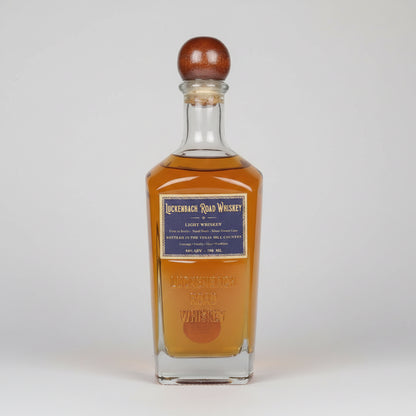 A bottle of Lickenbach Road Whiskey light whiskey with amber liquid, a dark blue label, and a brown wooden stopper, shown on a white background with a reflection.