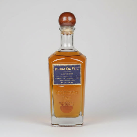 A bottle of Lickenbach Road Whiskey light whiskey with amber liquid, a dark blue label, and a brown wooden stopper, shown on a white background with a reflection.