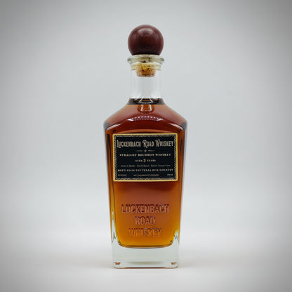 A bottle of Luckenbach Road Whiskey with amber-brown liquid, a black label with gold lettering, and a dark brown wooden stopper, shown on a plain white background.