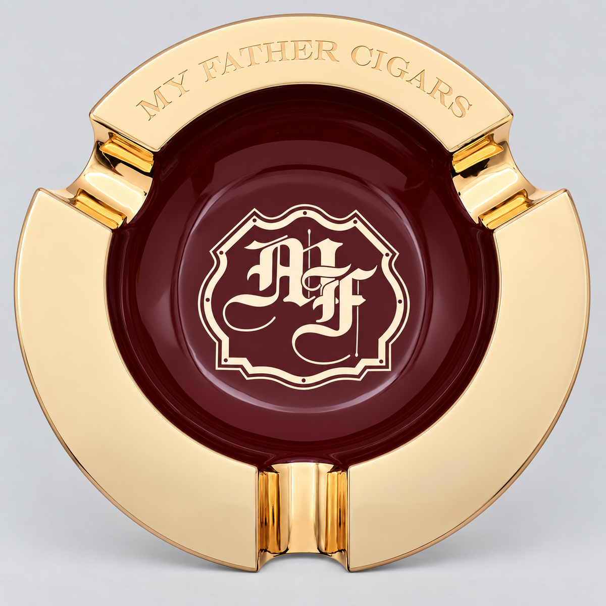A round gold and burgundy ashtray featuring a polished gold outer rim with the engraved text 'MY FATHER CIGARS' and a central burgundy circle with a gold monogram crest reading 'A F'.