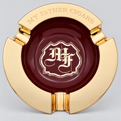 A round gold and burgundy ashtray featuring a polished gold outer rim with the engraved text 'MY FATHER CIGARS' and a central burgundy circle with a gold monogram crest reading 'A F'.