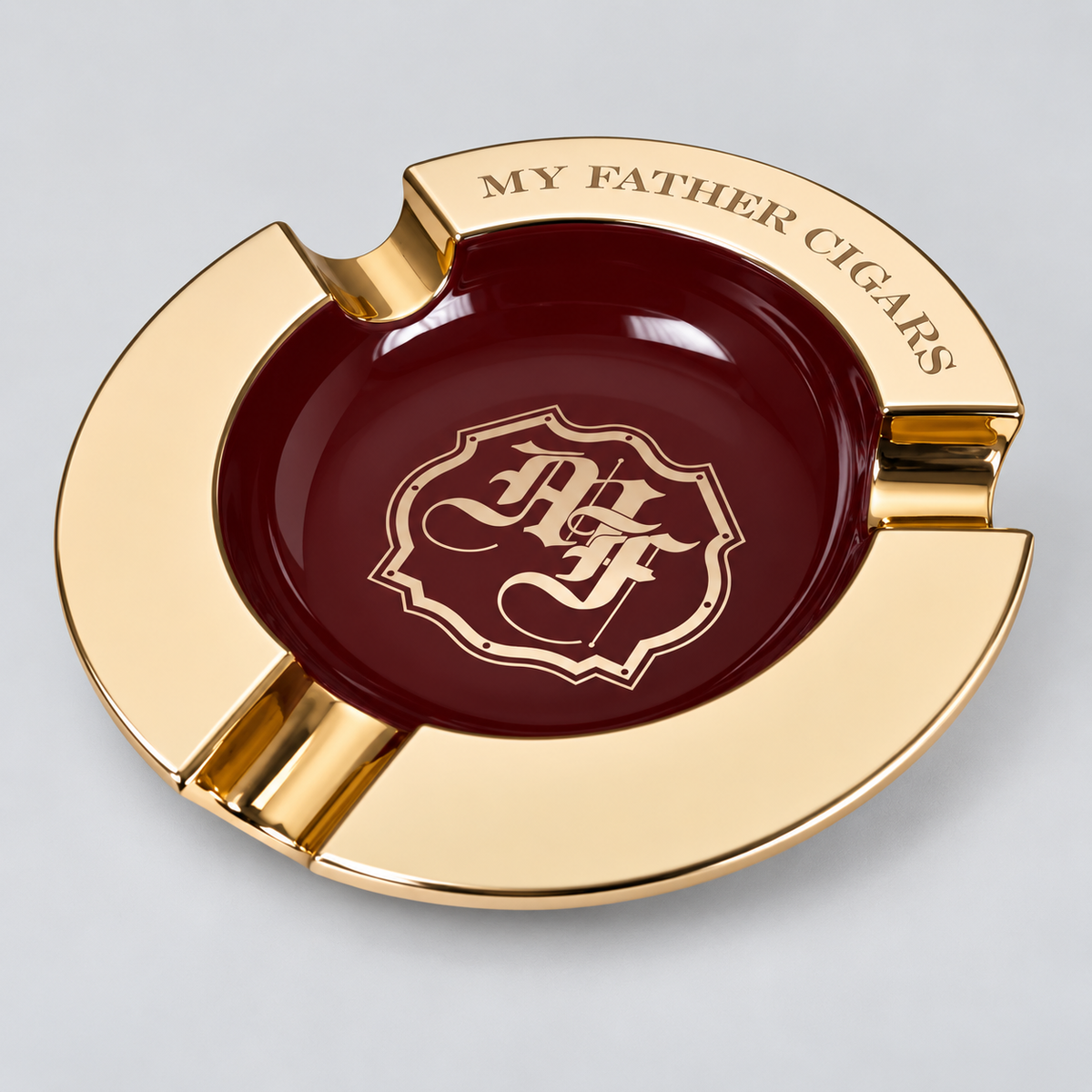 A luxury gold-plated circular cigar ashtray with a deep burgundy center. The outer rim is gold with the engraved text 'MY FATHER CIGARS' and a gold crest featuring the initials 'FC'.