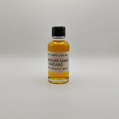 Bottle of Maker's Mark whiskey sample on a white surface with two silver bags in the background.