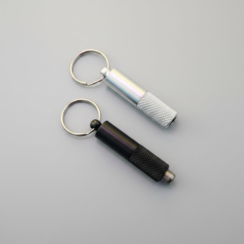 Two metal keychain tools on a wooden surface: a silver cylindrical tool with a knurled grip and a black cylindrical tool with a knurled grip, both attached to split rings.