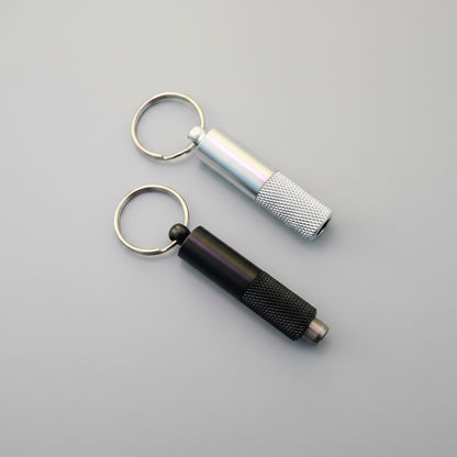 Two metal keychain tools on a wooden surface: a silver cylindrical tool with a knurled grip and a black cylindrical tool with a knurled grip, both attached to split rings.