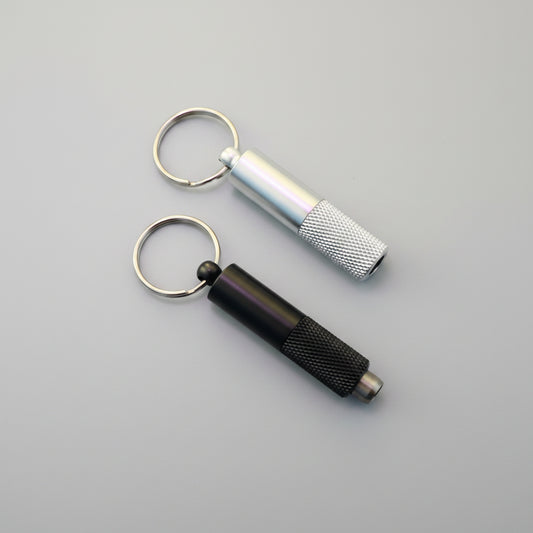 Two metal keychain tools on a wooden surface: a silver cylindrical tool with a knurled grip and a black cylindrical tool with a knurled grip, both attached to split rings.