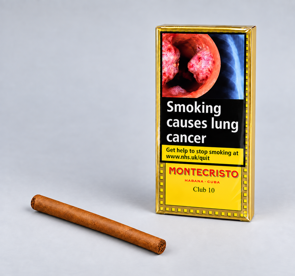 A single brown cigar lies on a light gray surface beside a gold-bordered Montecristo Club 10 pack with a smoking-health warning panel.