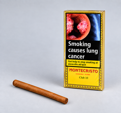 A single brown cigar lies on a light gray surface beside a gold-bordered Montecristo Club 10 pack with a smoking-health warning panel.