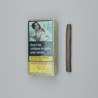 A Montecristo Marana Copa Mini 10 cigarette pack with a prominent smoking warning label, shown next to a single brown cigar on a solid taupe background.