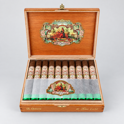 An open wooden cigar box displaying ten cigars inside, with a gray paper band across the front and a decorative interior lid featuring a colorful illustration. The brand name 'El Chaparral' is visible on the packaging.