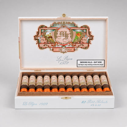 An open white cigar box with a decorative illustrated lid and multiple cigars arranged in a row inside. The cigars have cream wrappers with orange bands and dark brown fillings.