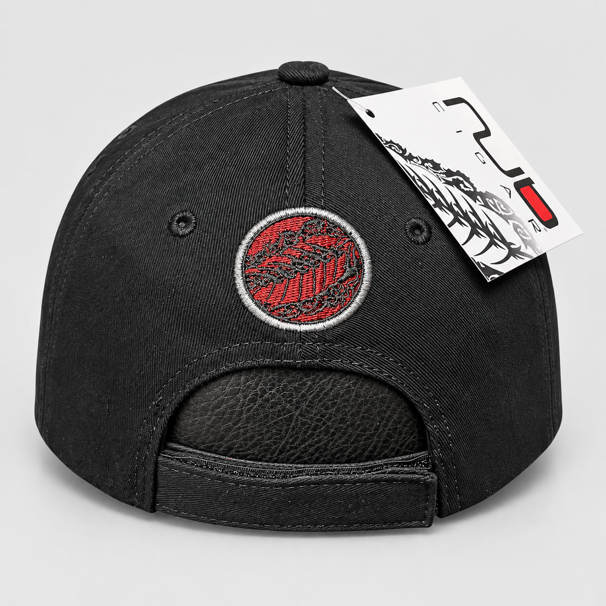 Black baseball cap with a red and black circular embroidered patch on the front, shown from the back with an adjustable strap and a white product tag attached, on a light gray background.