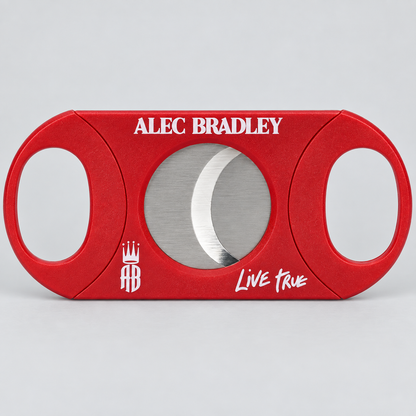 A red cigar cutter with a circular silver blade visible through the center, featuring 'ALEC BRADLEY' in white text at the top and 'Live True' with a crest logo at the bottom, set against a light gray background.