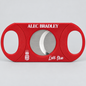 A red cigar cutter with a circular silver blade visible through the center, featuring 'ALEC BRADLEY' in white text at the top and 'Live True' with a crest logo at the bottom, set against a light gray background.