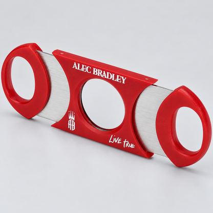 A red and silver guillotine-style cigar cutter with 'ALEC BRADLEY' and 'LIVE TRUE' printed on the blade, shown on a light gray background.