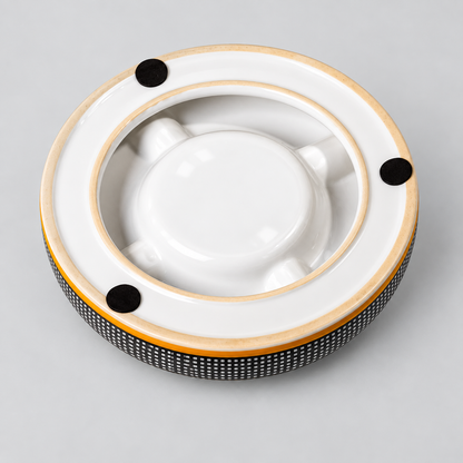 A round white ceramic ashtray with a black and white patterned lower band, a gold-colored rim, and three black circular feet, shown on a neutral gray background.
