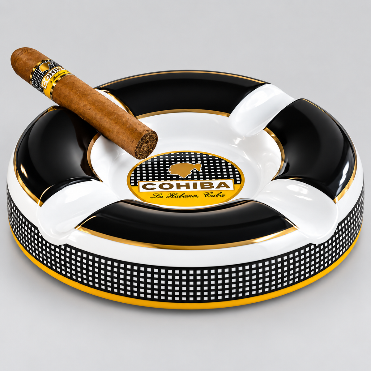 A round black, white, and gold ashtray with a cigar resting on top, featuring the COHIBA logo in the center, photographed on a light gray background.