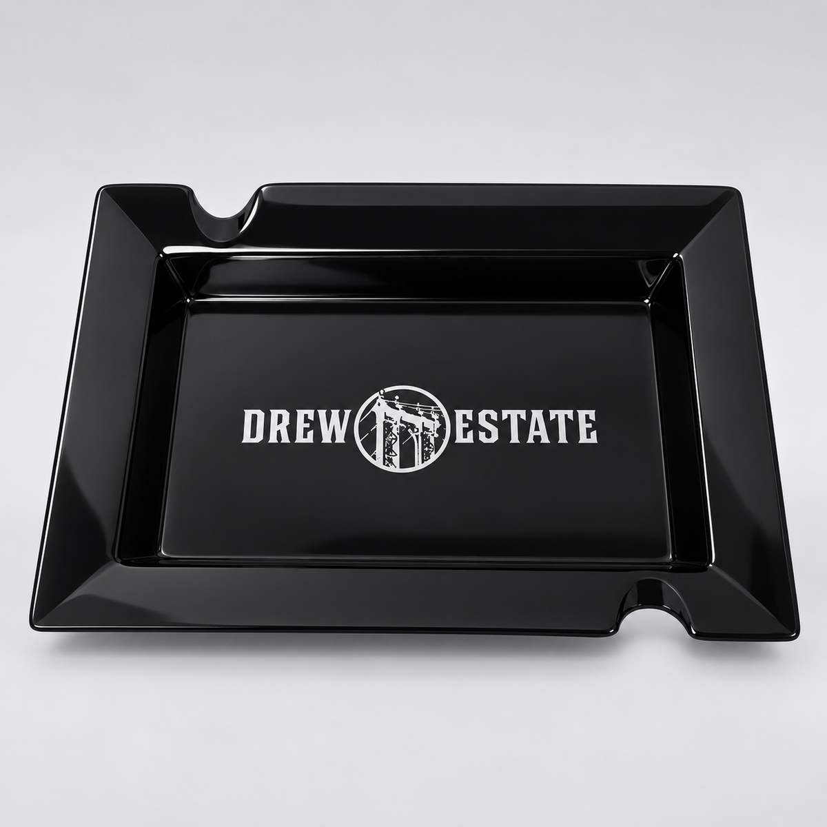 A glossy black rectangular tray with two small cutouts at the top corners and a white 'DREW ESTATE' logo printed in the center, shown on a light gray background.