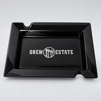 A glossy black rectangular tray with two small cutouts at the top corners and a white 'DREW ESTATE' logo printed in the center, shown on a light gray background.