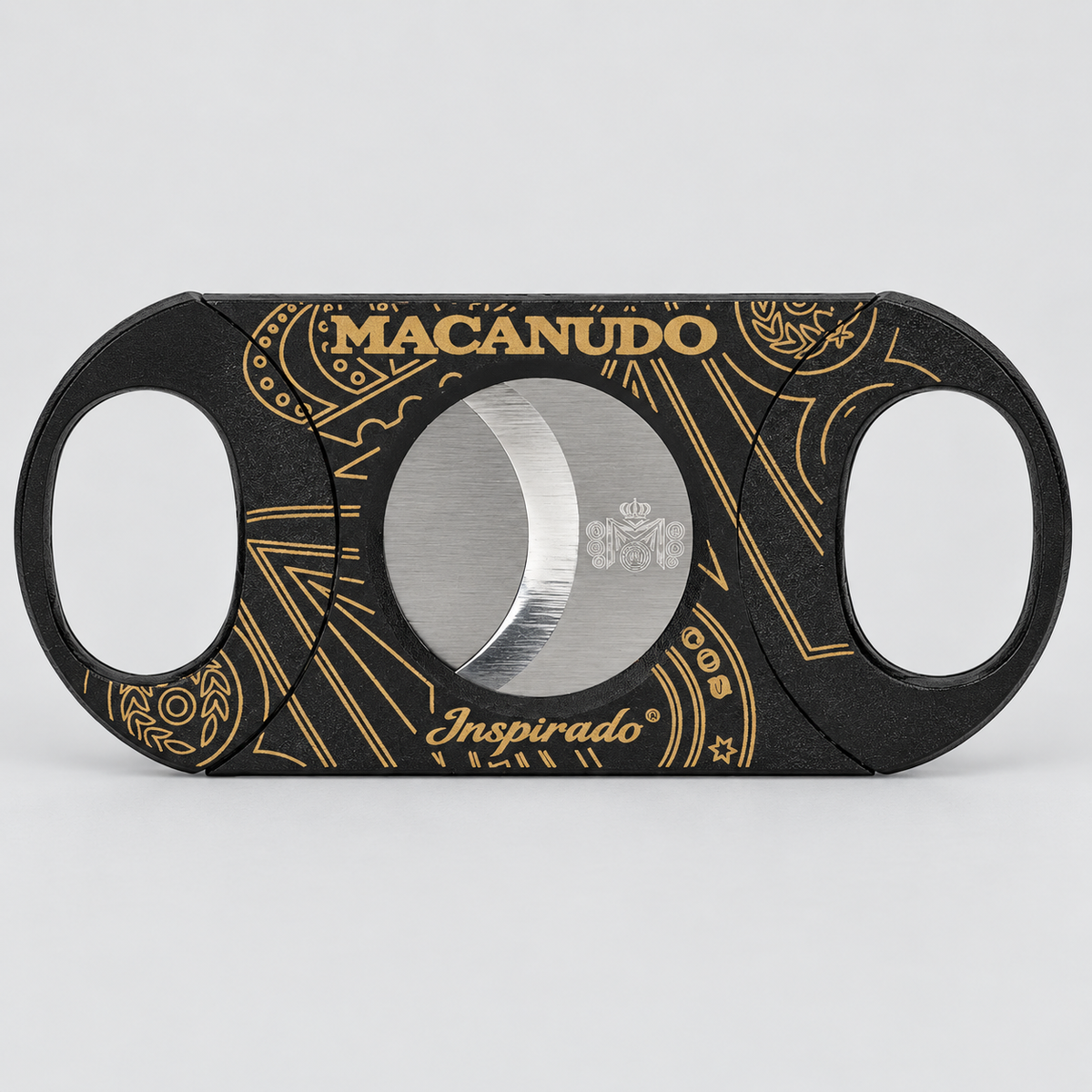 A black cigar cutter with gold ornamental patterns and the brand name 'MACANUDO' in gold on the top, and 'Inspirado' on the bottom, shown on a light gray background.