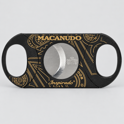 A black cigar cutter with gold ornamental patterns and the brand name 'MACANUDO' in gold on the top, and 'Inspirado' on the bottom, shown on a light gray background.