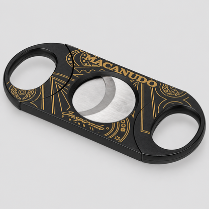 A black cigar cutter with gold decorative patterns and the brand name 'MACANUDO' on a light gray background.