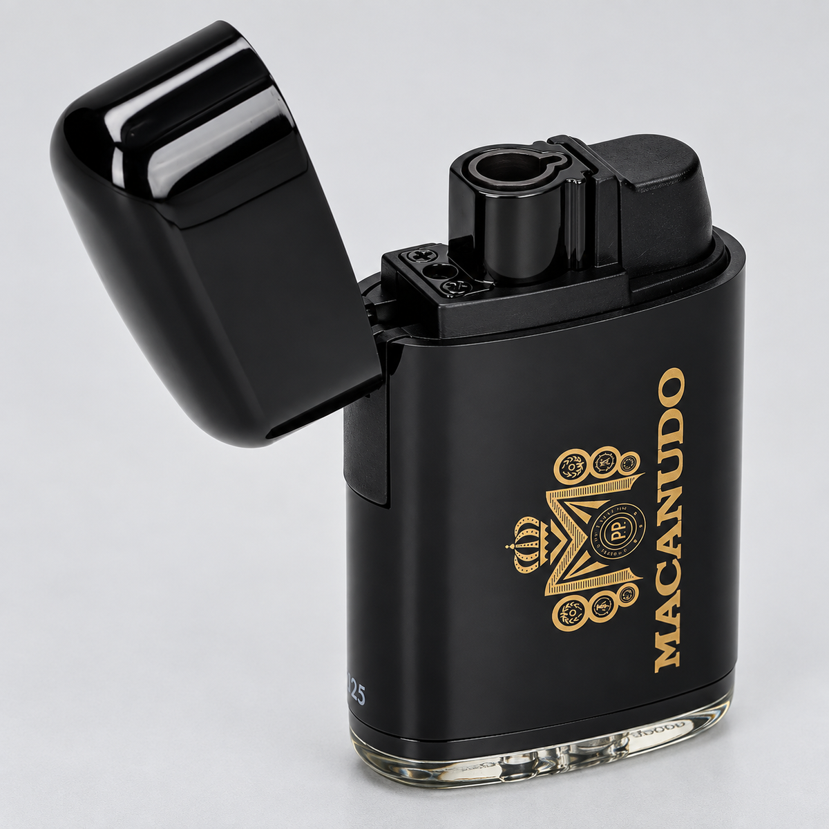 A black metallic lighter with its lid open, featuring a gold crown and circular emblem on the front and the brand name 'MACANUDO' printed vertically in gold. The lighter has a clear glass base and is photographed on a light gray background.