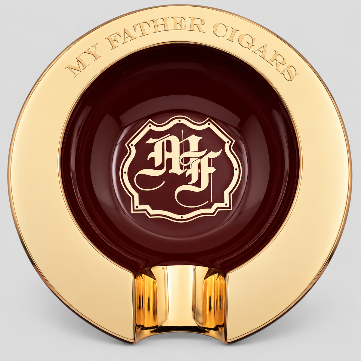 A luxury circular ashtray with a polished gold outer rim engraved with 'MY FATHER CIGARS' and a central dark burgundy recessed area featuring a gold monogram crest. The ashtray is shown on a neutral light gray background.
